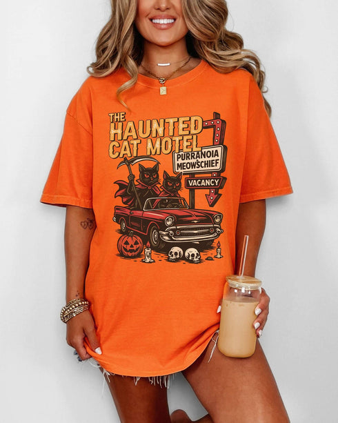 Woman wearing a burnt-orange-colored 'The Haunted Cat Motel' shirt with a funny retro Halloween graphic of two cats driving to a spooky motel.