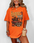 Woman wearing a burnt-orange-colored 'The Haunted Cat Motel' shirt with a funny retro Halloween graphic of two cats driving to a spooky motel.