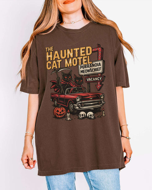 Woman wearing an espresso-colored 'The Haunted Cat Motel' shirt with a funny retro Halloween graphic of two cats driving to a spooky motel.