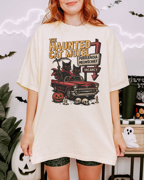 Woman wearing an ivory-colored 'The Haunted Cat Motel' shirt with a funny retro Halloween graphic of two cats driving to a spooky motel.