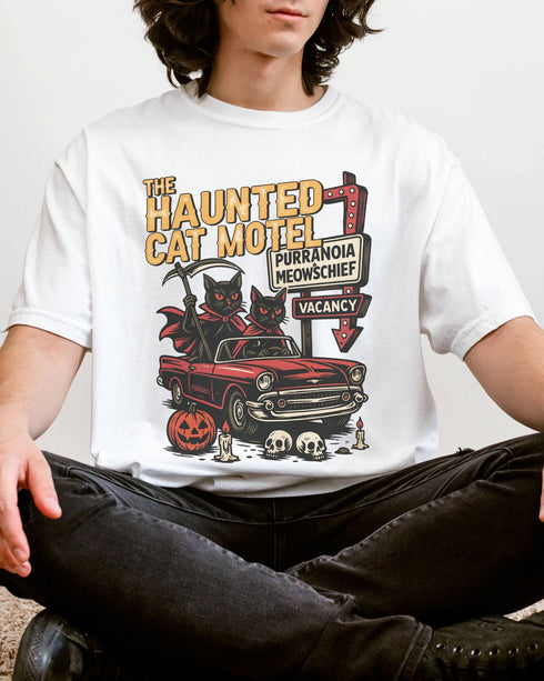 Man wearing a white-colored 'The Haunted Cat Motel' shirt with a funny retro Halloween graphic of two cats driving to a spooky motel.