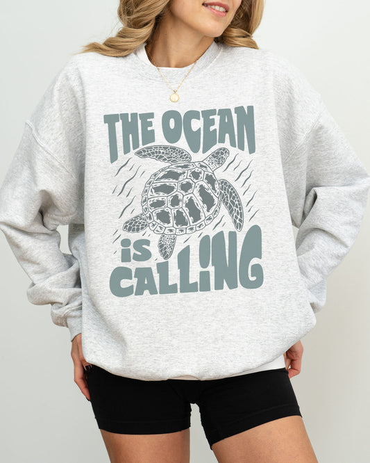 Model wearing ash color sweatshirt with 'The Ocean Is Calling' graphic design