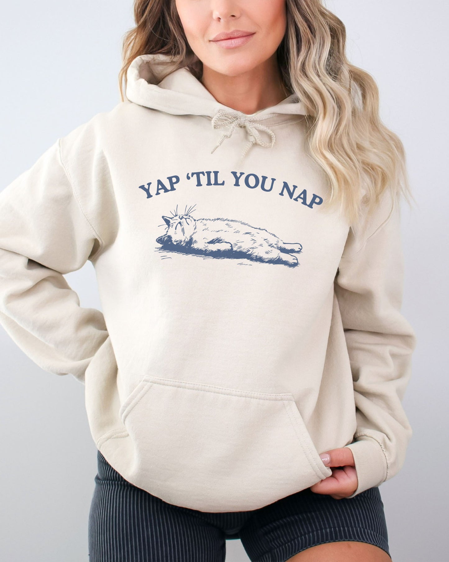 Woman wearing a sand beige hoodie featuring a funny retro sleeping cat graphic and the quote 'Yap 'Til You Nap'.