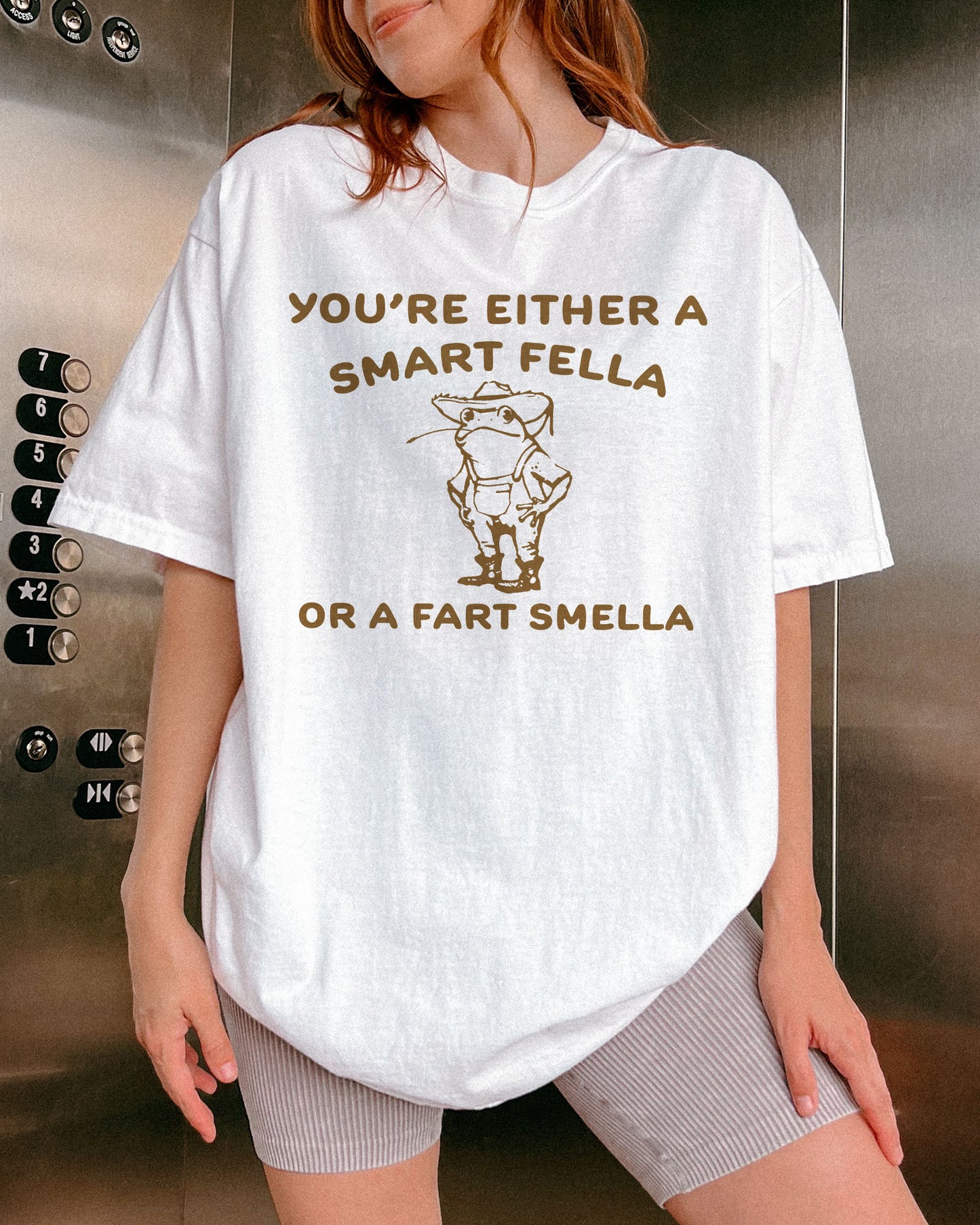 Woman wearing a white "You’re Either a Smart Fella or a Fart Smella" shirt with a funny retro outline graphic of a cowboy frog.