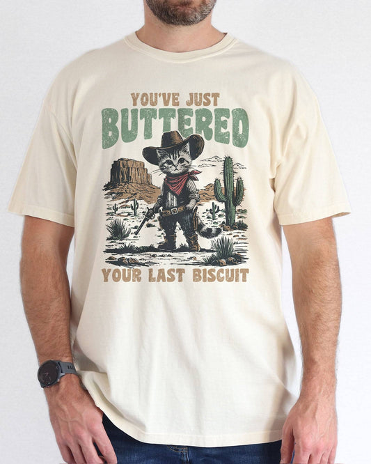 Model wearing natural color shirt with 'You've Just Buttered Your Last Biscuit' cowboy kitten graphic design