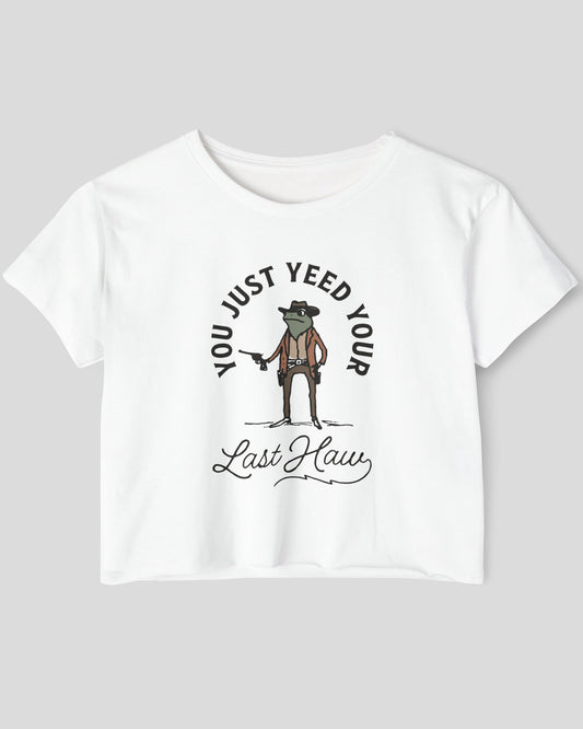 Flat lay of a white crop top with a funny retro cowboy sheriff frog graphic and the text 'You Just Yeed Your Last Haw'.