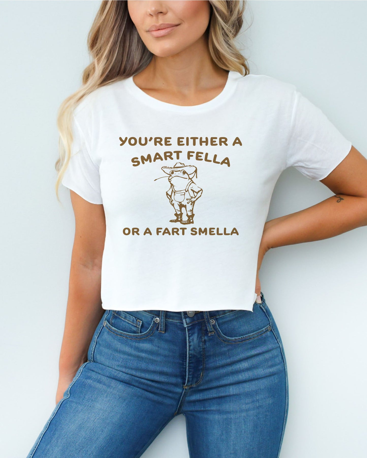 Woman wearing white-colored 'You're Either A Smart Fella Or A Fart Smella' crop top featuring a funny retro cowboy frog graphic.