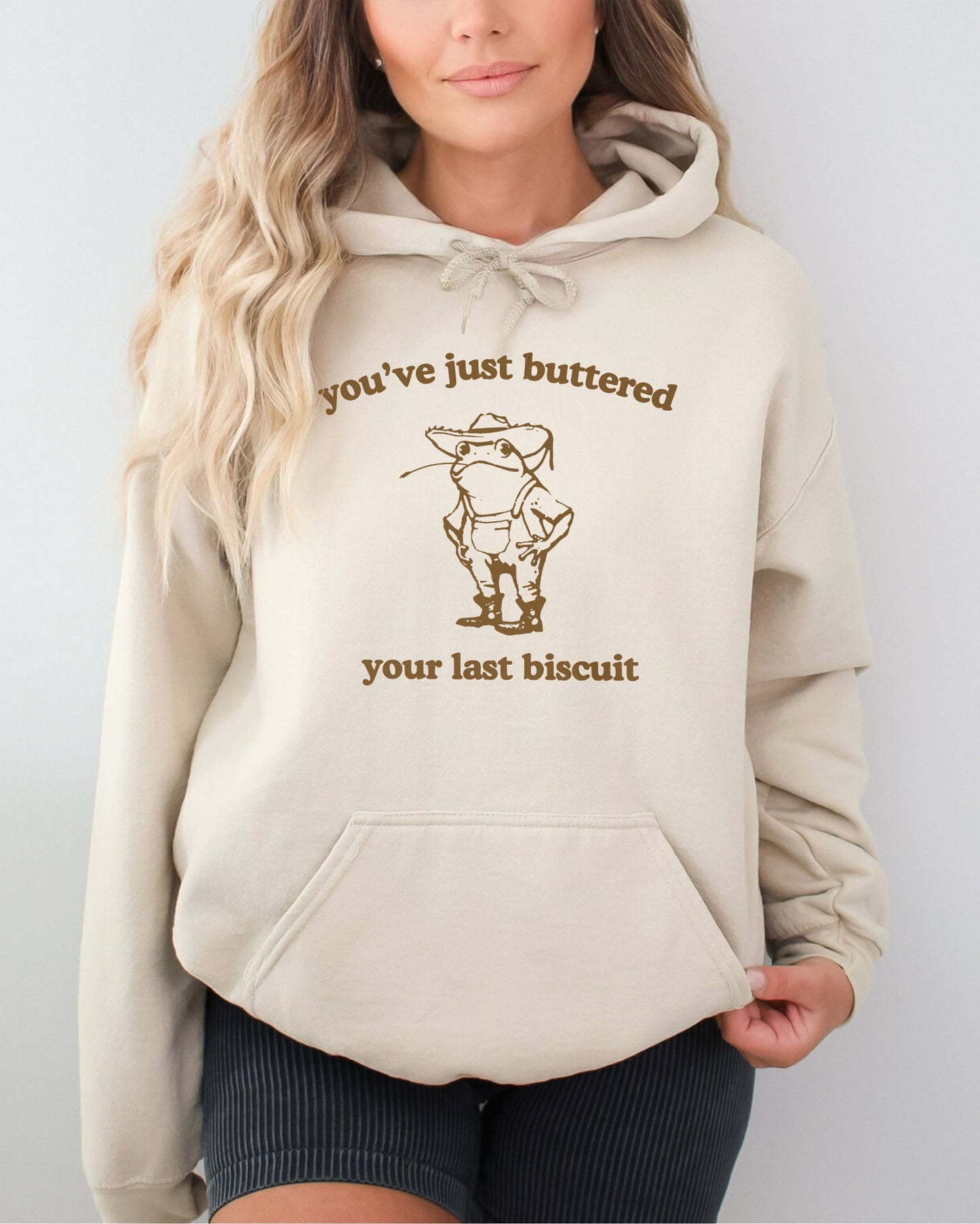 Woman in a sand beige hoodie featuring a vintage country frog graphic with the quote 'You've Just Buttered Your Last Biscuit'.