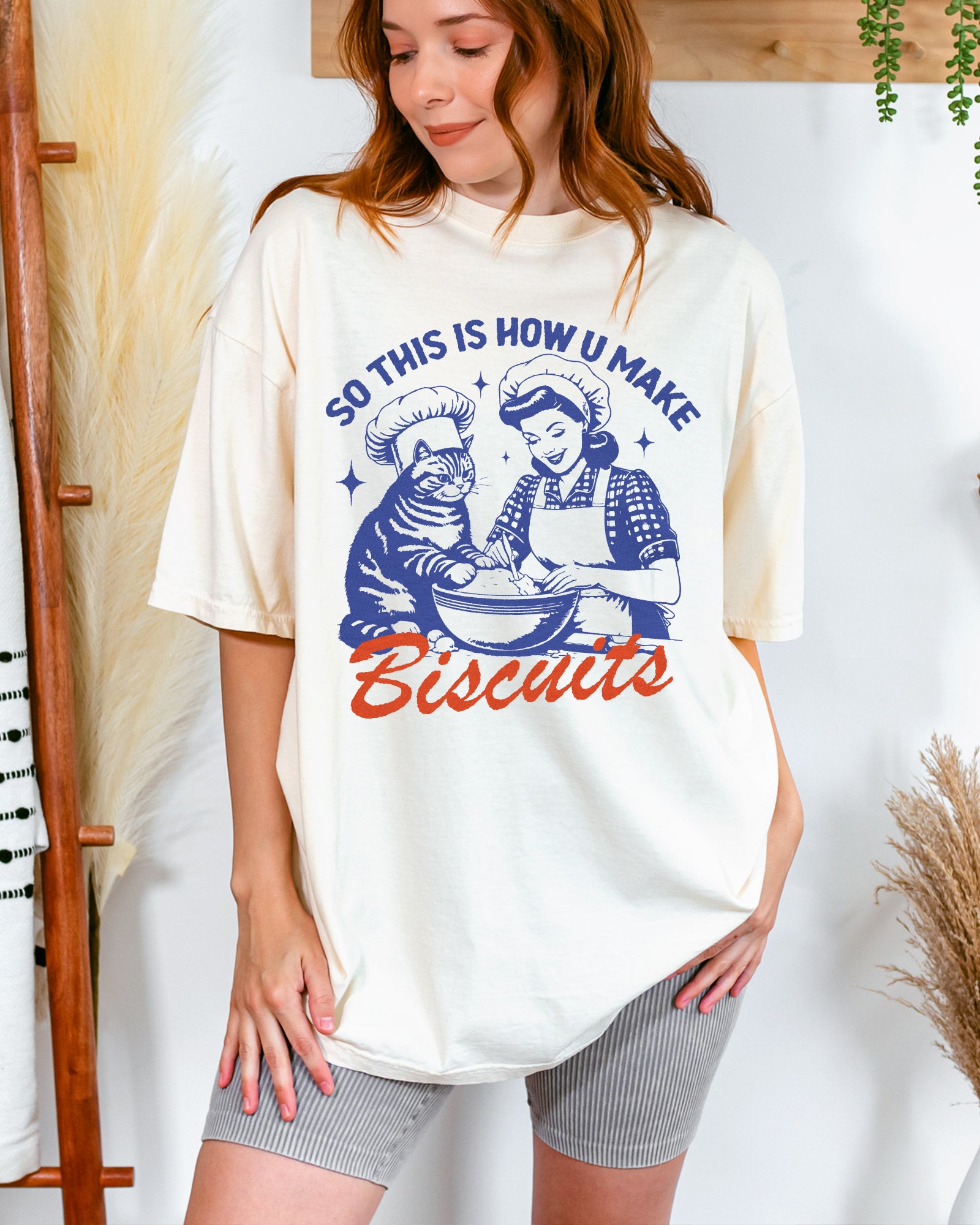 Woman wearing "So This Is How You Make Biscuits" shirt in ivory with funny retro graphic of cat and woman making biscuits together