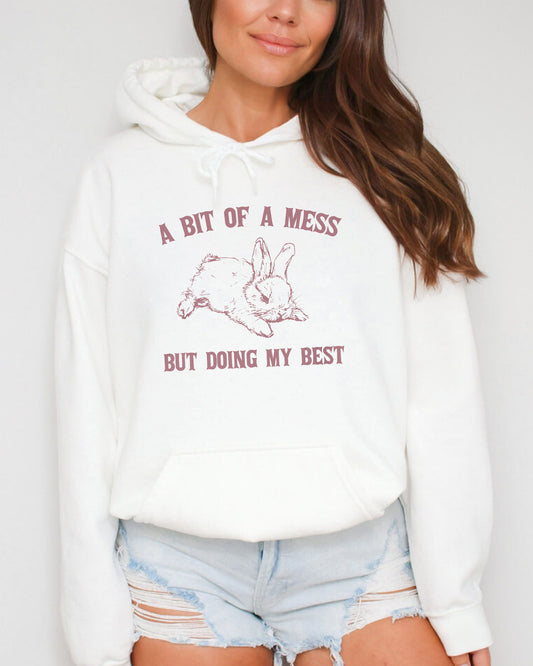 Woman wearing a white hoodie with the phrase 'A Bit Of A Mess But Doing My Best' and a retro bunny line art graphic.