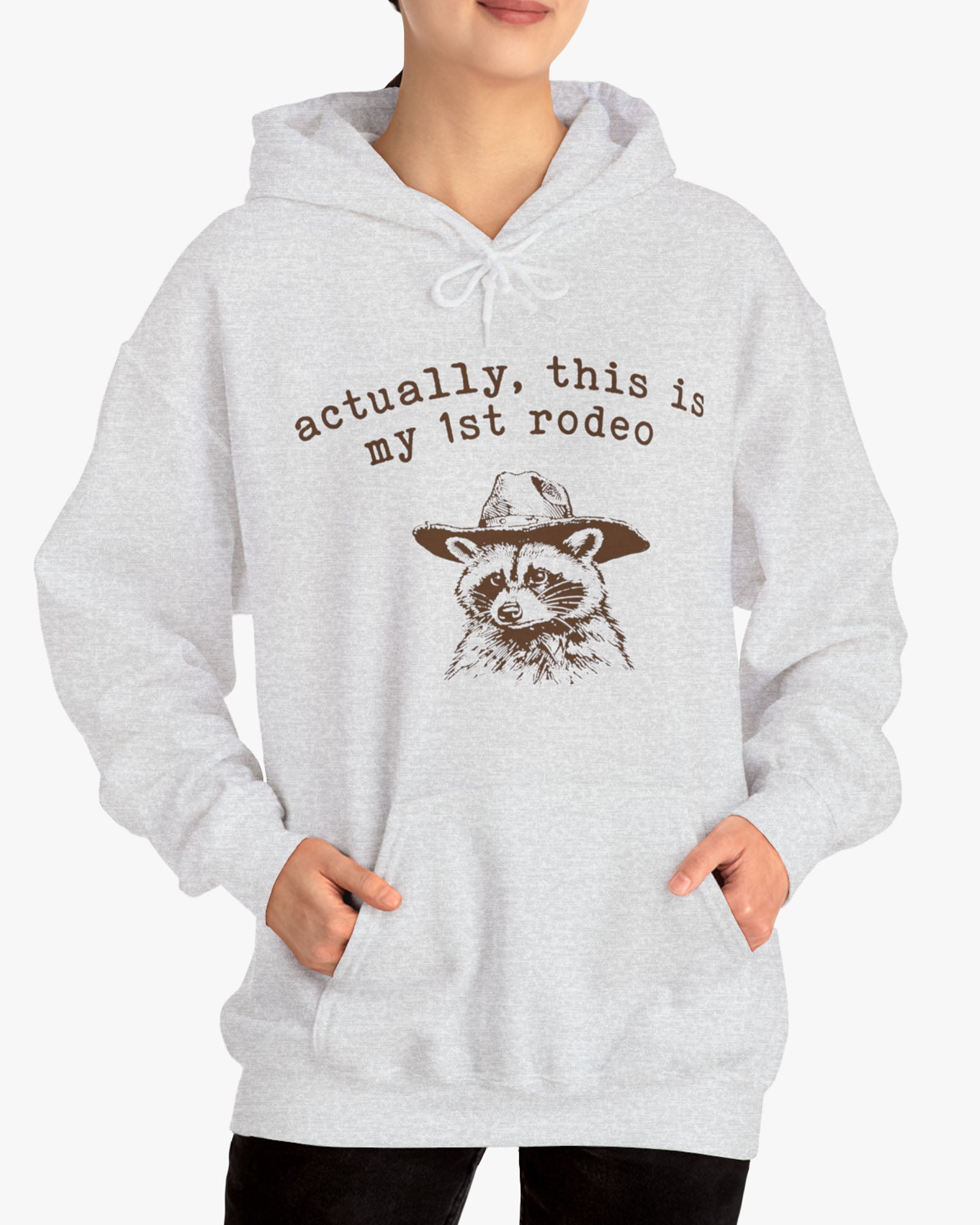 Woman wearing an 'Actually This is My First Rodeo' hoodie in ash gray, featuring a retro western graphic design and unisex fit.