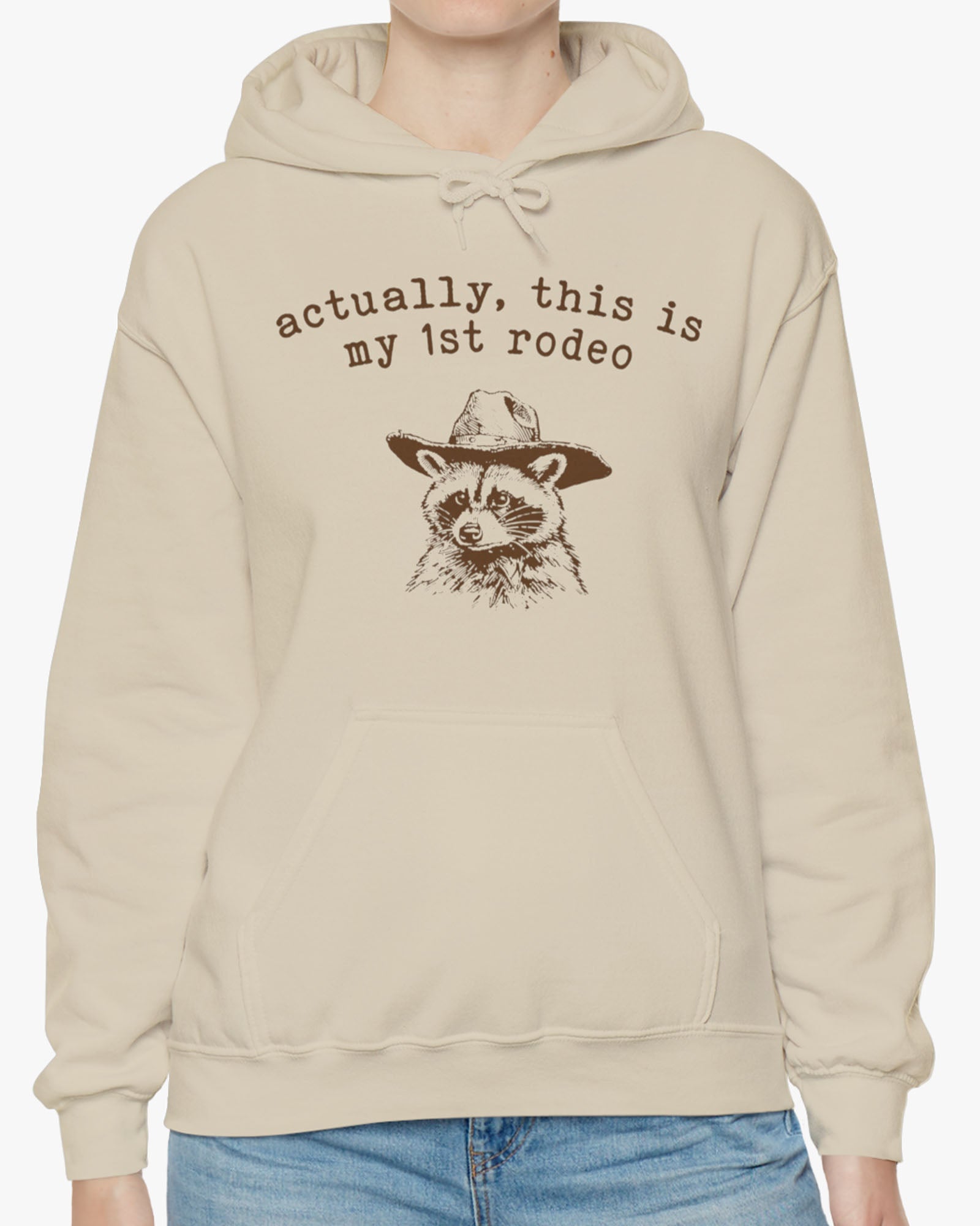 Woman wearing an 'Actually This is My First Rodeo' hoodie in sand beige, featuring a retro western graphic design and unisex fit.