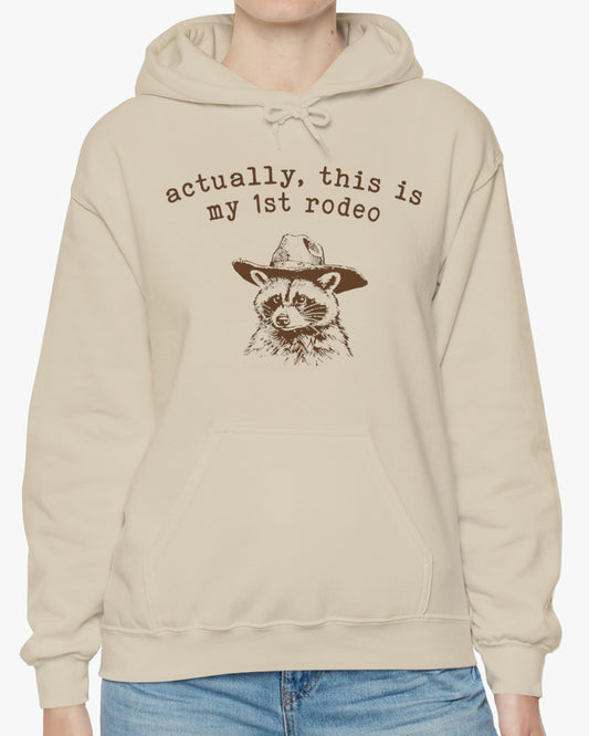 Woman wearing an 'Actually This is My First Rodeo' hoodie in sand beige, featuring a retro western graphic design and unisex fit.