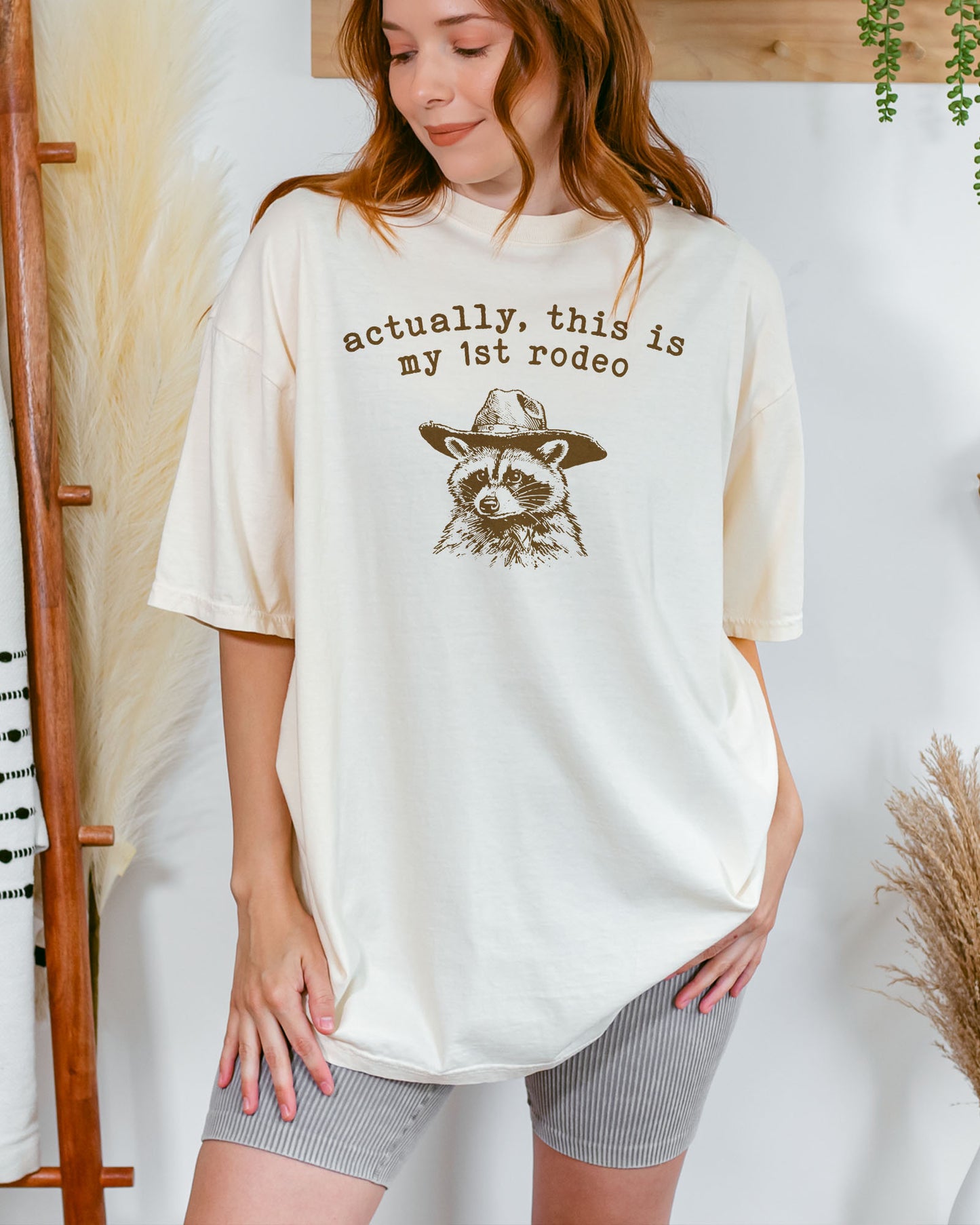 Woman wearing an ivory-colored 'Actually This Is My First Rodeo' shirt with a funny retro cowboy raccoon.