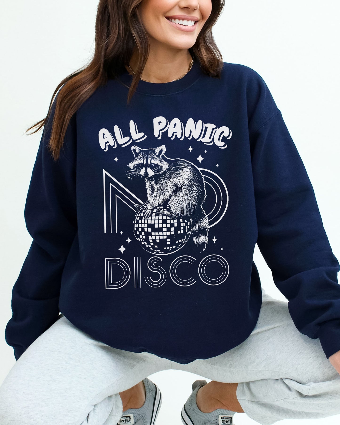 Woman wearing a navy blue sweatshirt featuring a funny retro raccoon sitting on a disco ball graphic and the quote 'All Panic No Disco'.