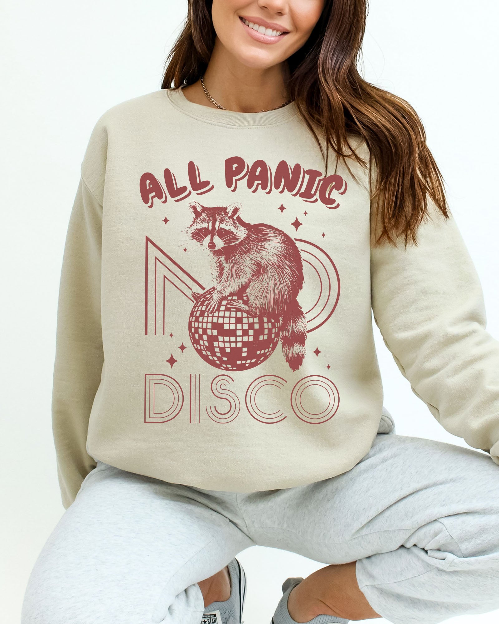 Woman wearing a sand beige sweatshirt featuring a funny retro raccoon sitting on a disco ball graphic and the quote 'All Panic No Disco'.