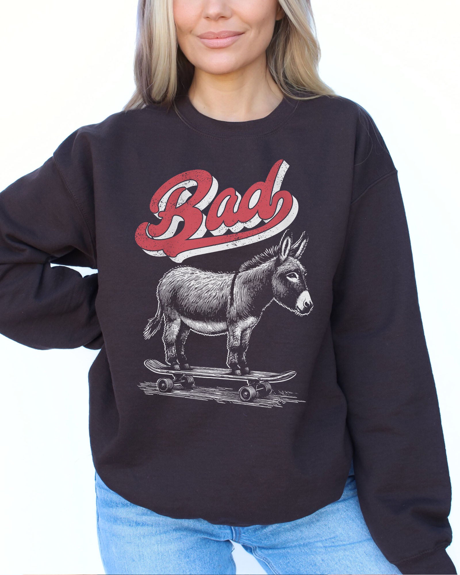 Woman wearing a dark chocolate color sweatshirt with a retro graphic of a donkey riding a skateboard and the word 'Bad' above it.
