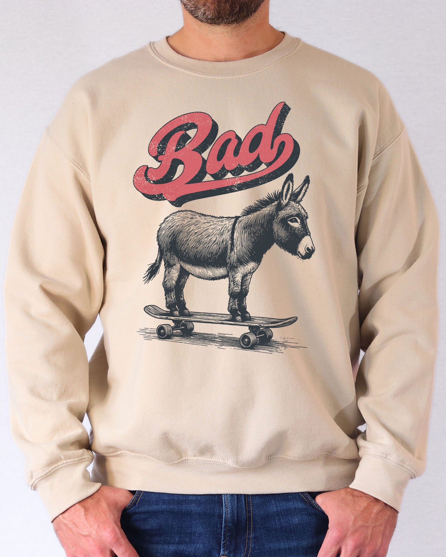 Man wearing a sand beige sweatshirt with a retro graphic of a donkey riding a skateboard and the word 'Bad' above it.