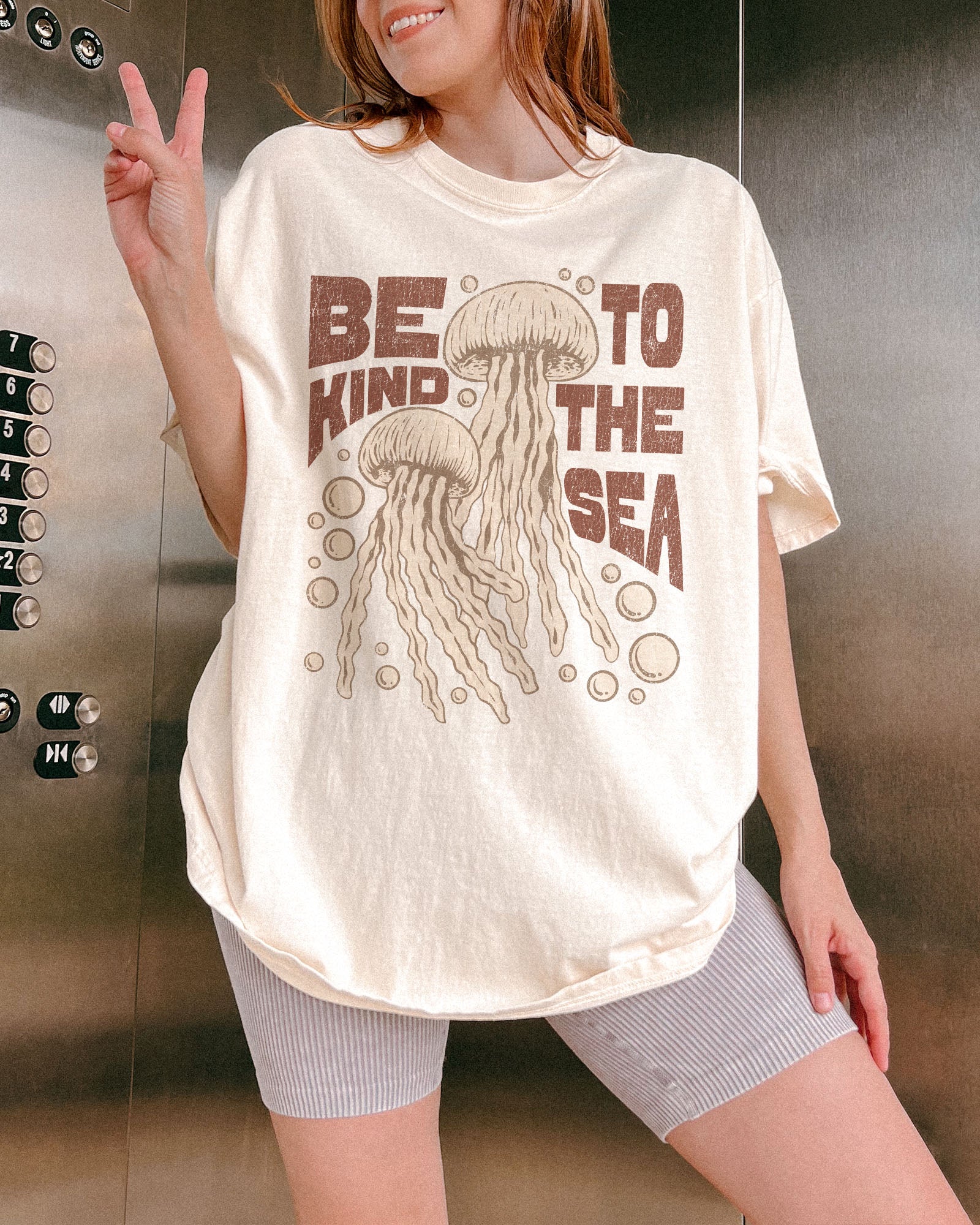 Woman wearing an ivory "Be Kind To The Sea" shirt with a retro-style graphic of two jellyfish floating underwater.
