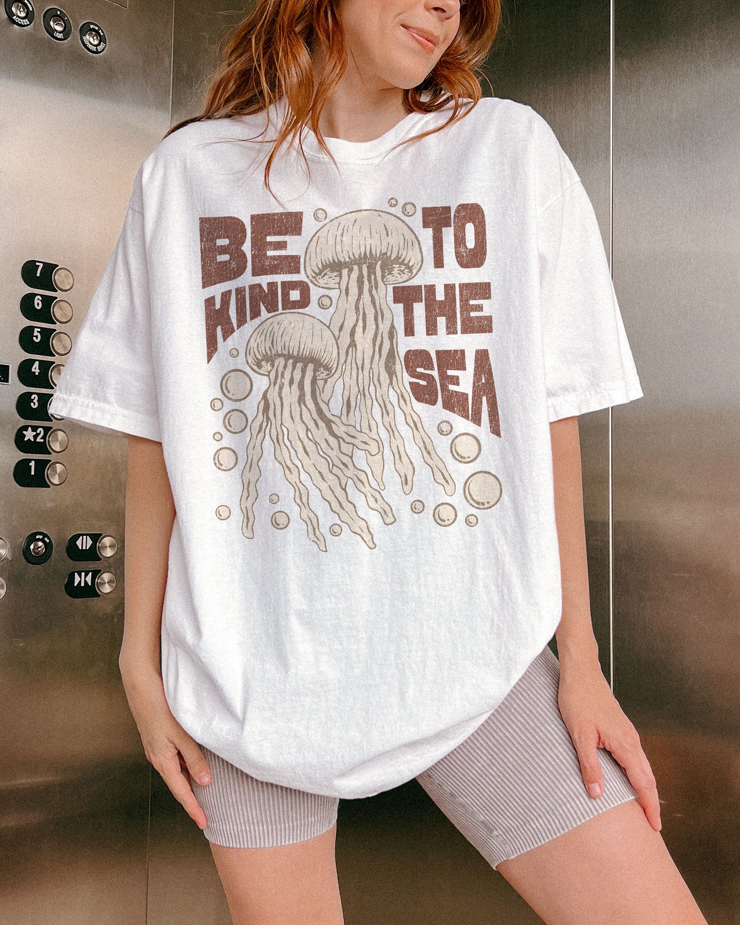 Woman wearing a white "Be Kind To The Sea" shirt with a retro-style graphic of two jellyfish floating underwater.