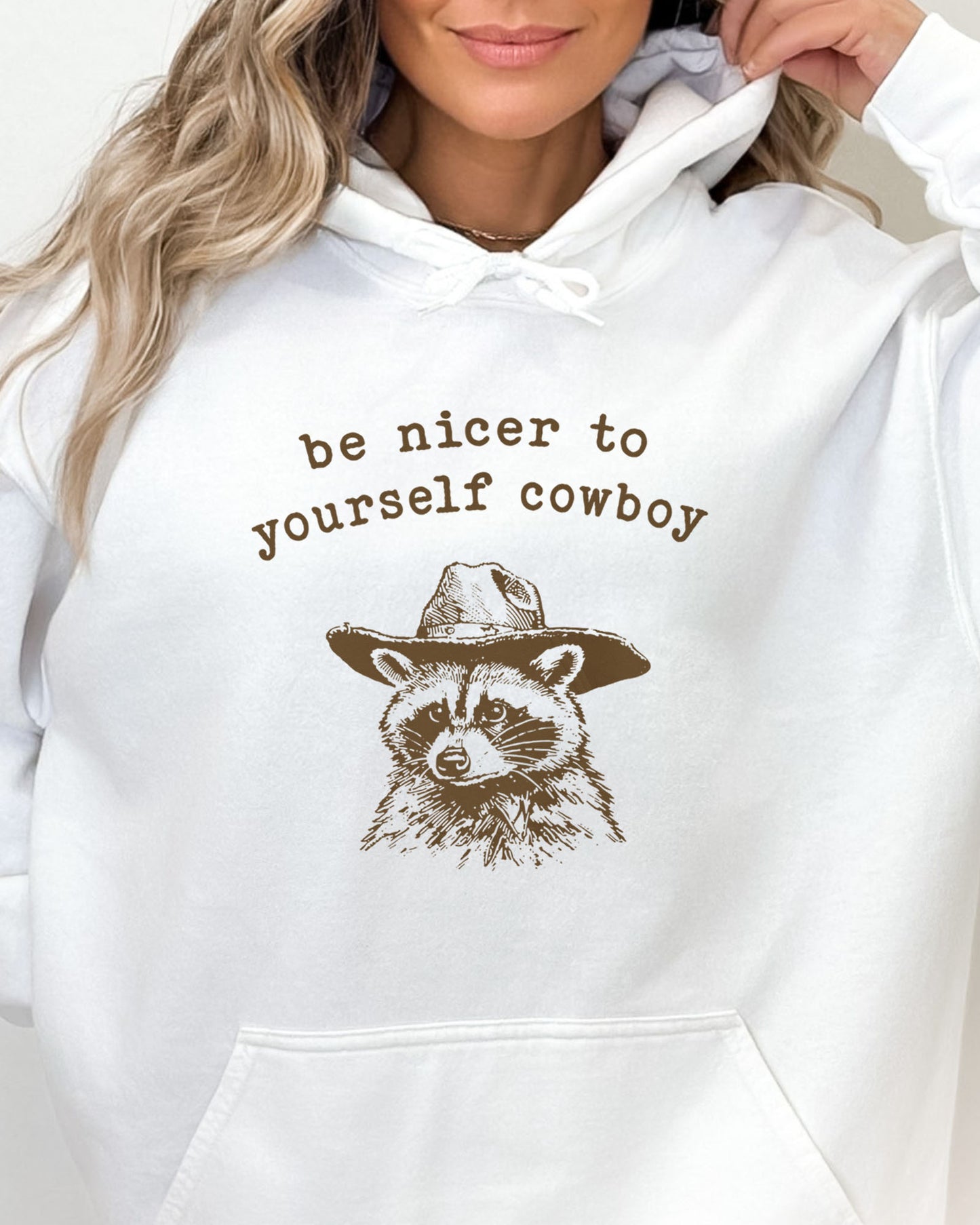 Close-up of a woman wearing a white hoodie featuring a funny retro cowboy raccoon graphic and the quote 'Be Nicer To Yourself Cowboy'.
