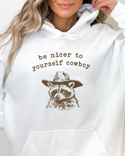 Close-up of a woman wearing a white hoodie featuring a funny retro cowboy raccoon graphic and the quote 'Be Nicer To Yourself Cowboy'.