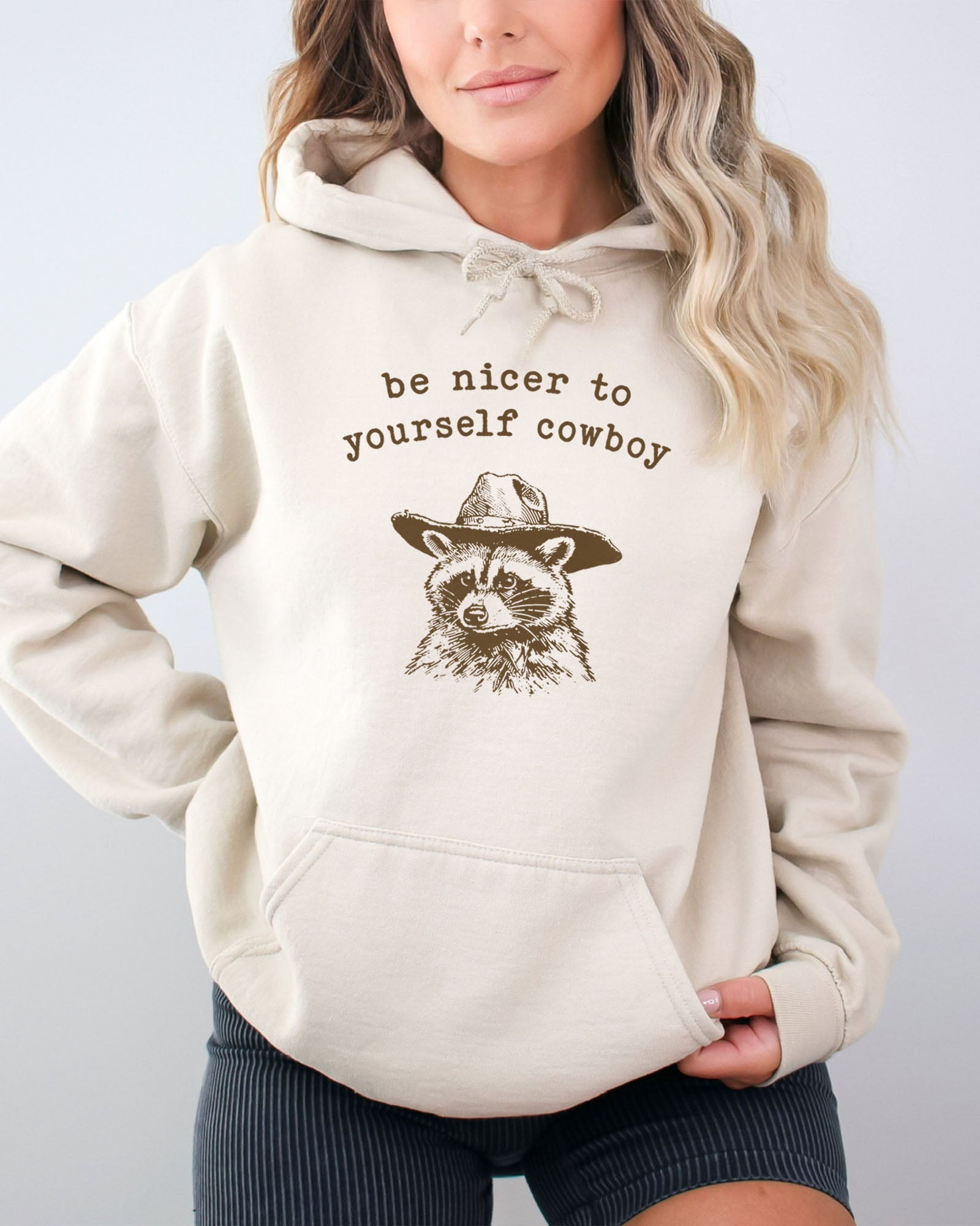 Woman wearing a sand beige hoodie featuring a funny retro cowboy raccoon graphic and the quote 'Be Nicer To Yourself Cowboy'.