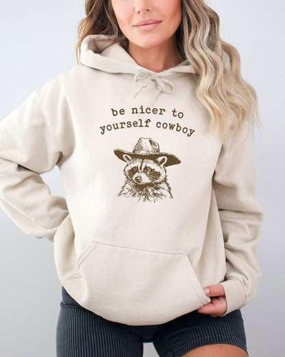 Woman wearing a sand beige hoodie featuring a funny retro cowboy raccoon graphic and the quote 'Be Nicer To Yourself Cowboy'.