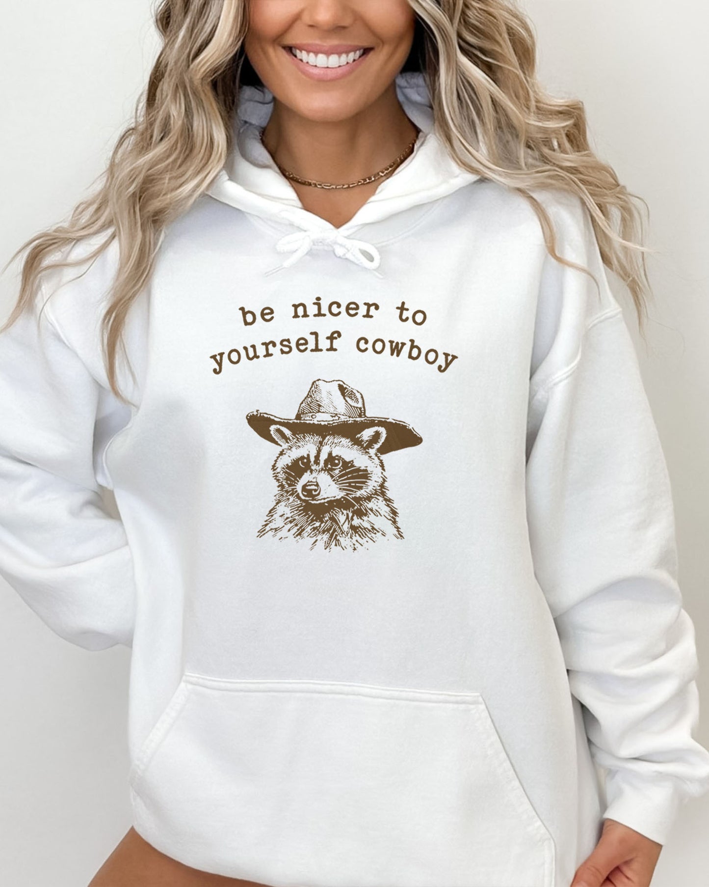 Woman wearing a white hoodie featuring a funny retro cowboy raccoon graphic and the quote 'Be Nicer To Yourself Cowboy'.