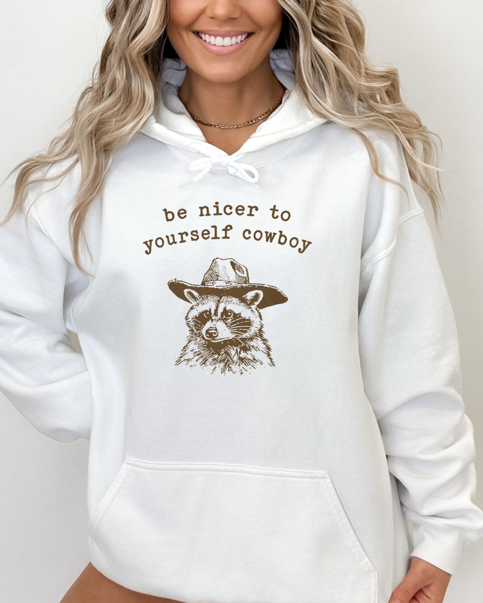 Woman wearing a white hoodie featuring a funny retro cowboy raccoon graphic and the quote 'Be Nicer To Yourself Cowboy'.