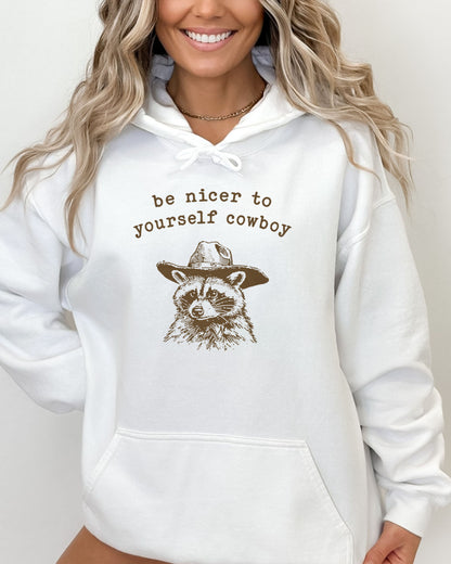 Woman wearing a white hoodie featuring a funny retro cowboy raccoon graphic and the quote 'Be Nicer To Yourself Cowboy'.