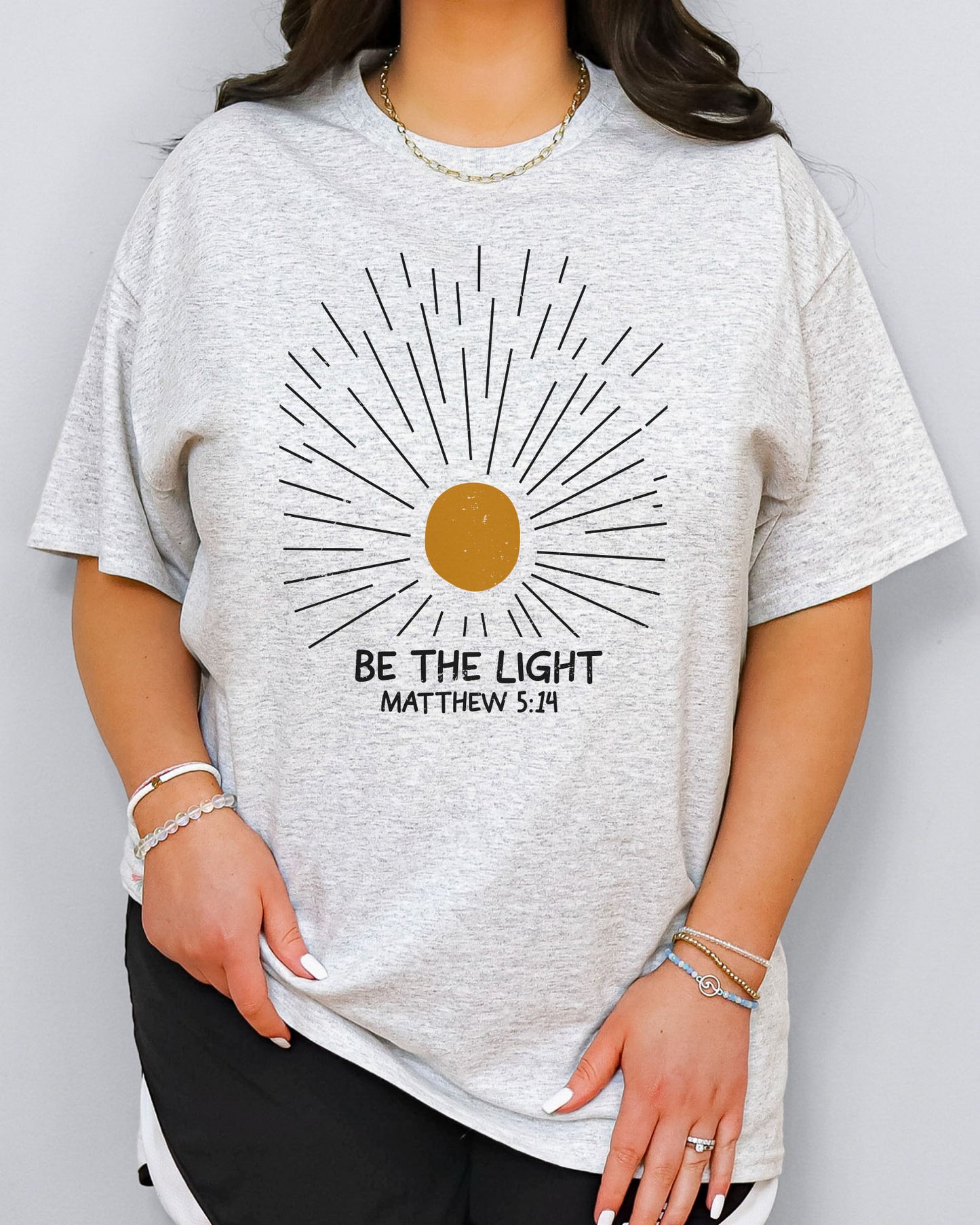 Model wearing ash color shirt with 'Be The Light - Matthew 5:14' graphic design