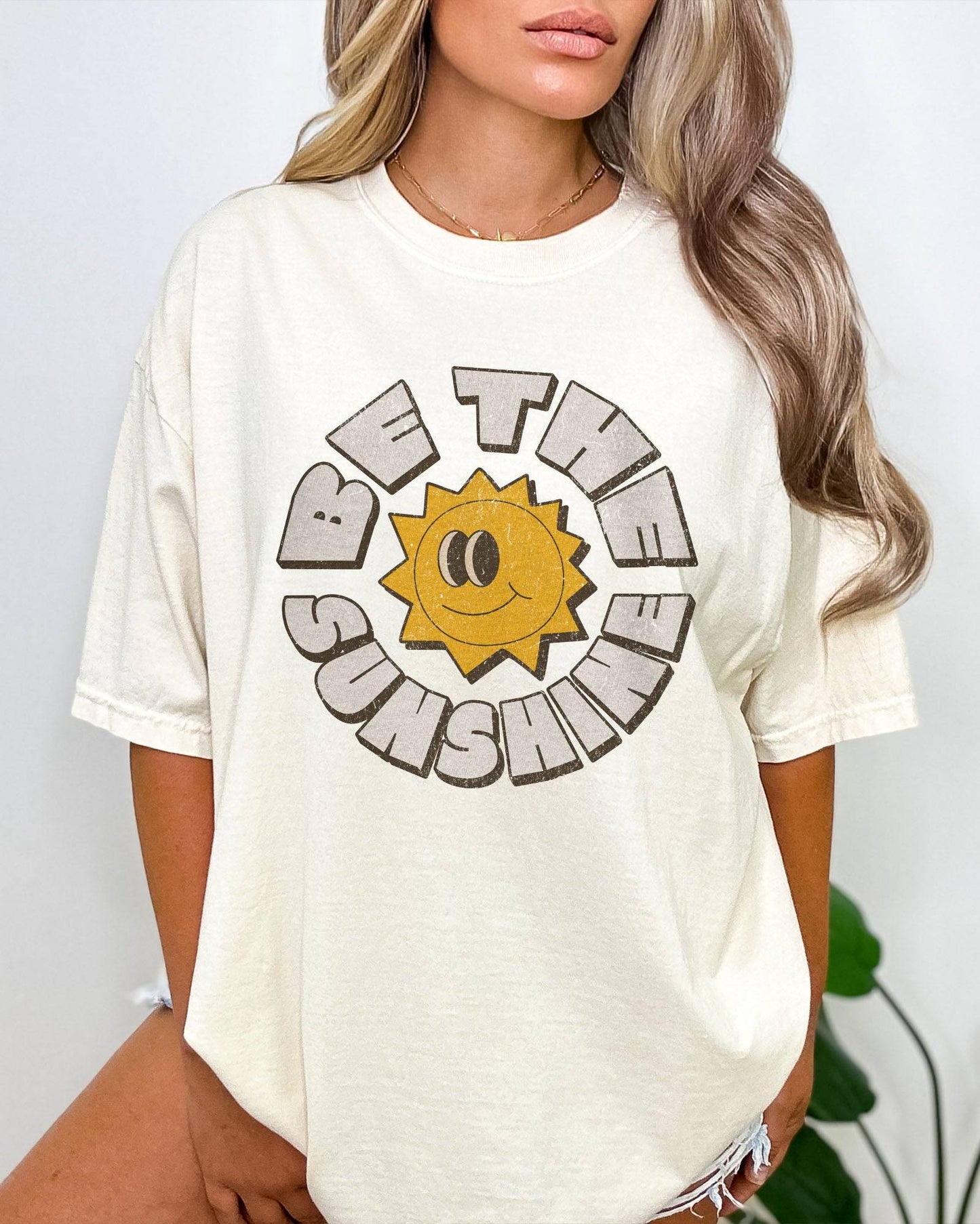 Woman wearing an ivory-colored Be The Sunshine shirt with a retro boho sun graphic.