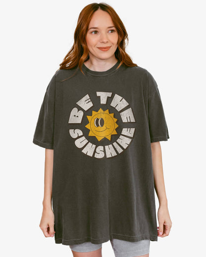 Woman wearing a pepper Be The Sunshine shirt with a retro boho sun graphic.
