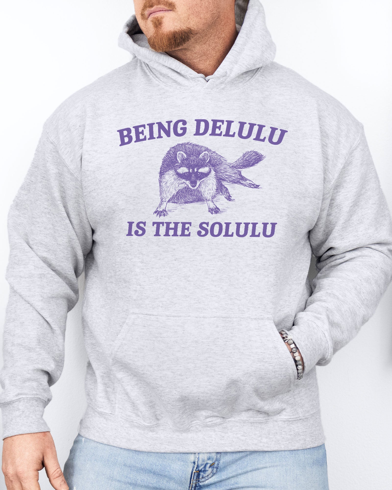 Man wearing an ash gray hoodie featuring a unny retro-style fat raccoon graphic and the quote 'Being Delulu Is The Solulu'. 
