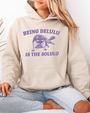Woman wearing a sand beige hoodie featuring a unny retro-style fat raccoon graphic and the quote 'Being Delulu Is The Solulu'. 