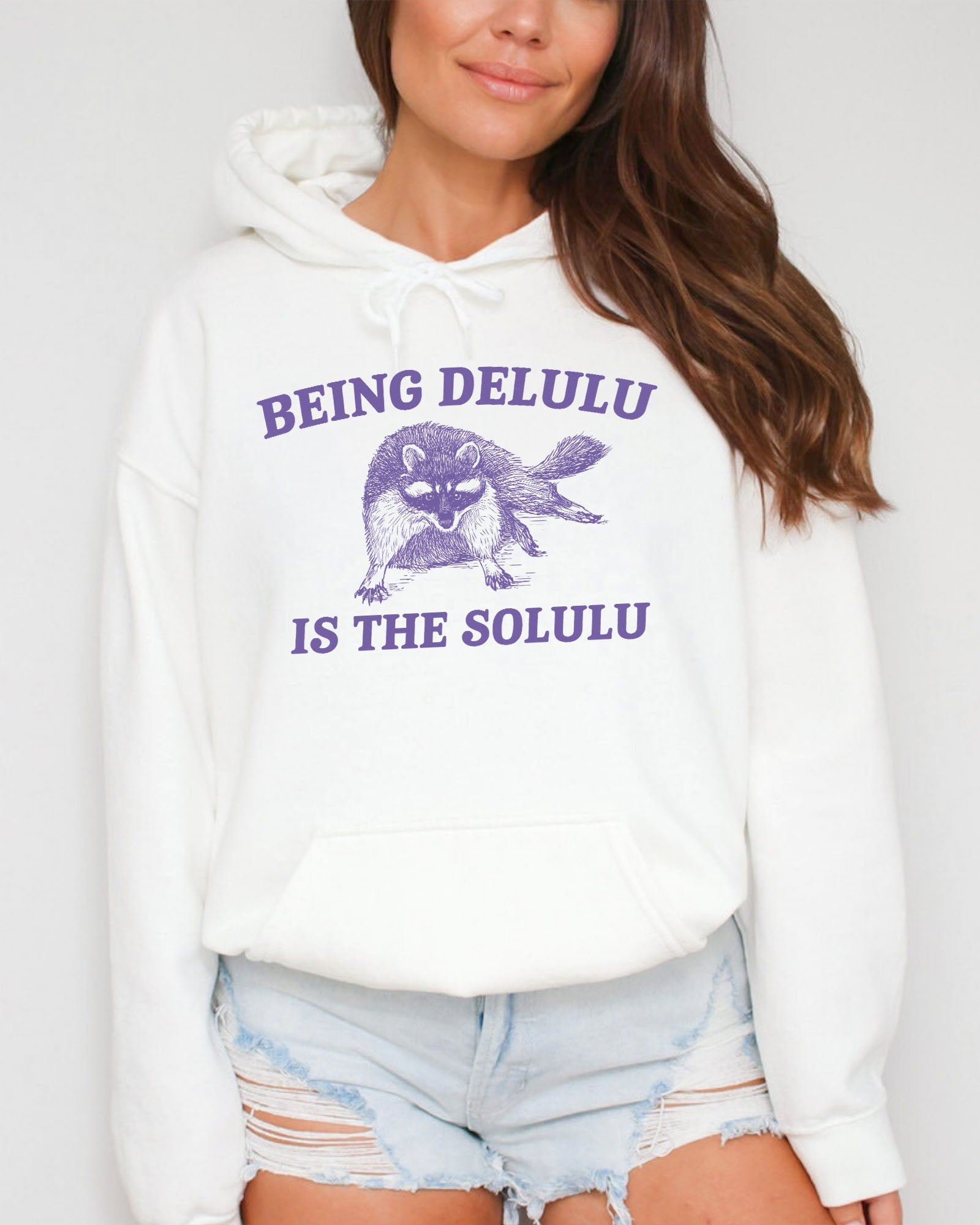 Woman wearing a white hoodie featuring a unny retro-style fat raccoon graphic and the quote 'Being Delulu Is The Solulu'. 