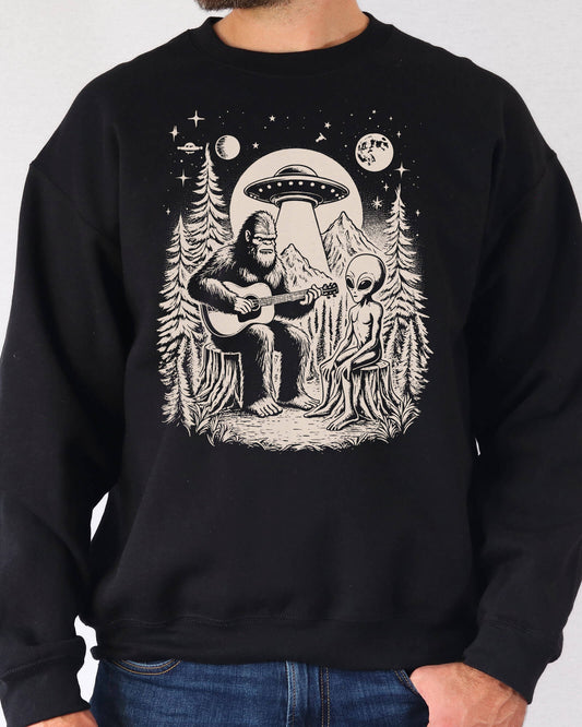 Man wearing a black color sweatshirt featuring a retro graphic of Bigfoot playing guitar with an Alien And UFO.