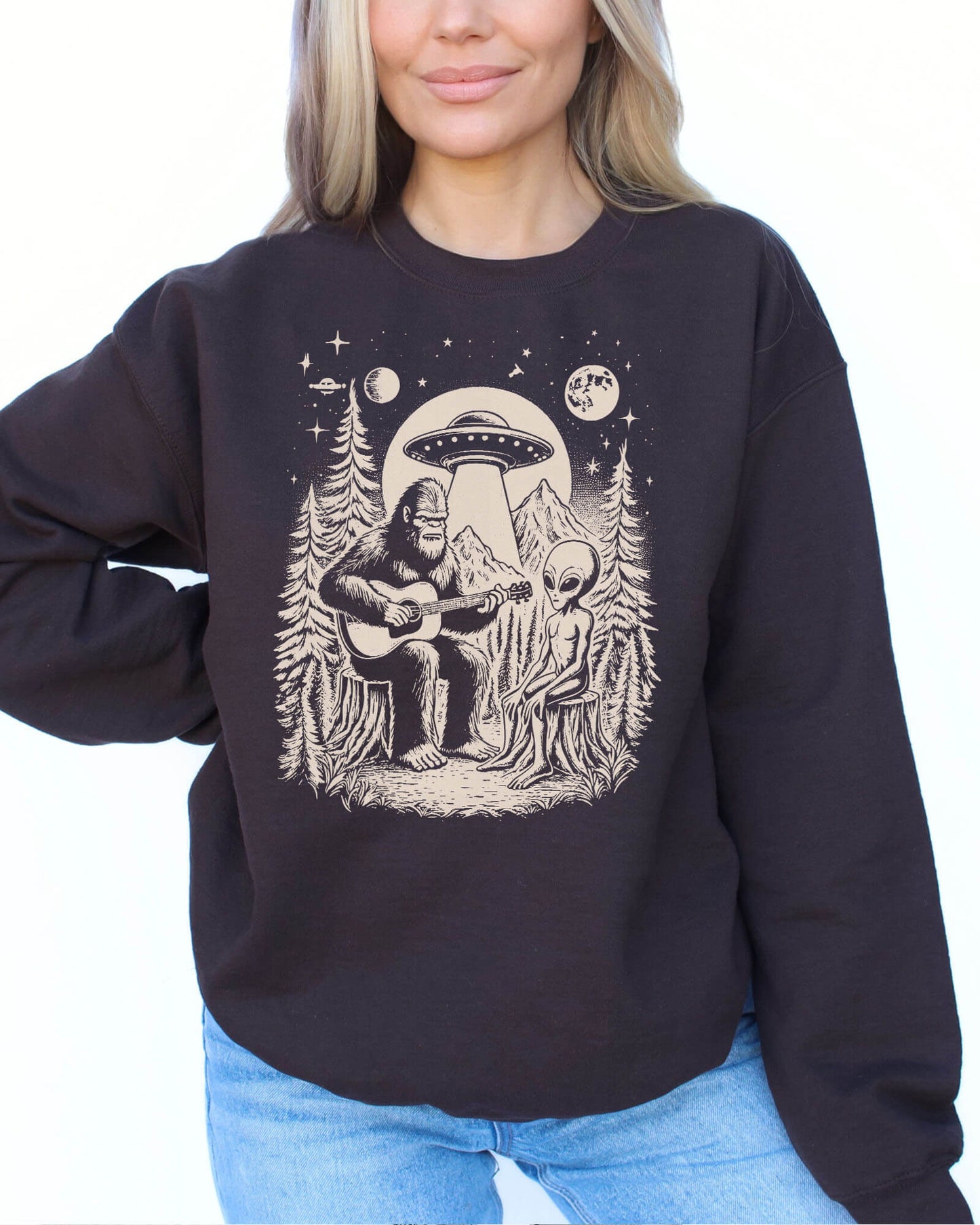 Woman wearing a dark chocolate color sweatshirt featuring a retro graphic of Bigfoot playing guitar with an alien and UFO.