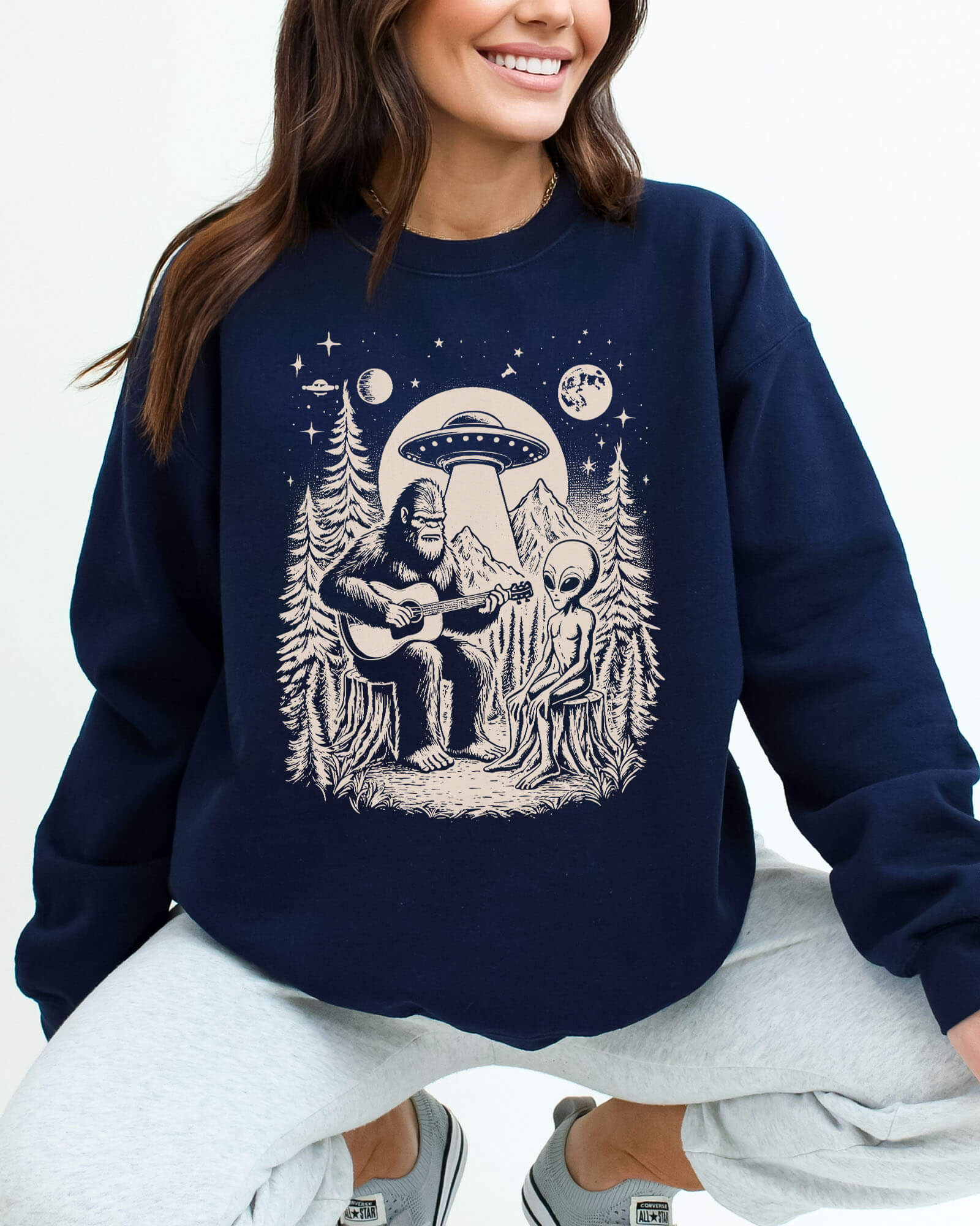 Woman wearing a navy blue sweatshirt featuring a retro graphic of Bigfoot playing guitar with an alien And UFO.