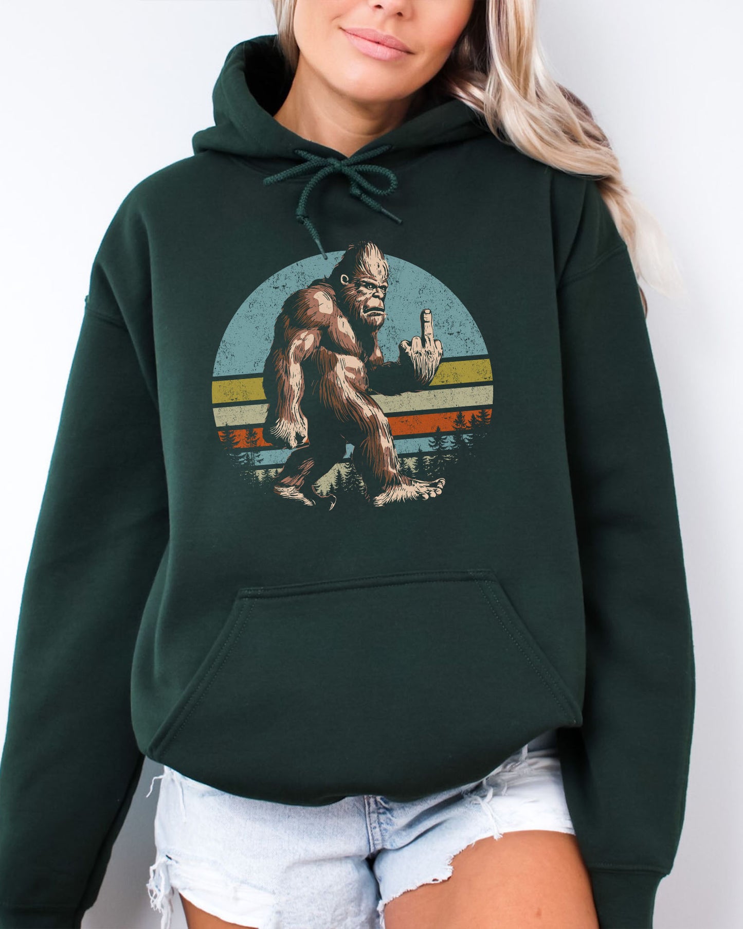 Woman wearing a forest green hoodie with a funny retro graphic of Bigfoot showing the middle finger.