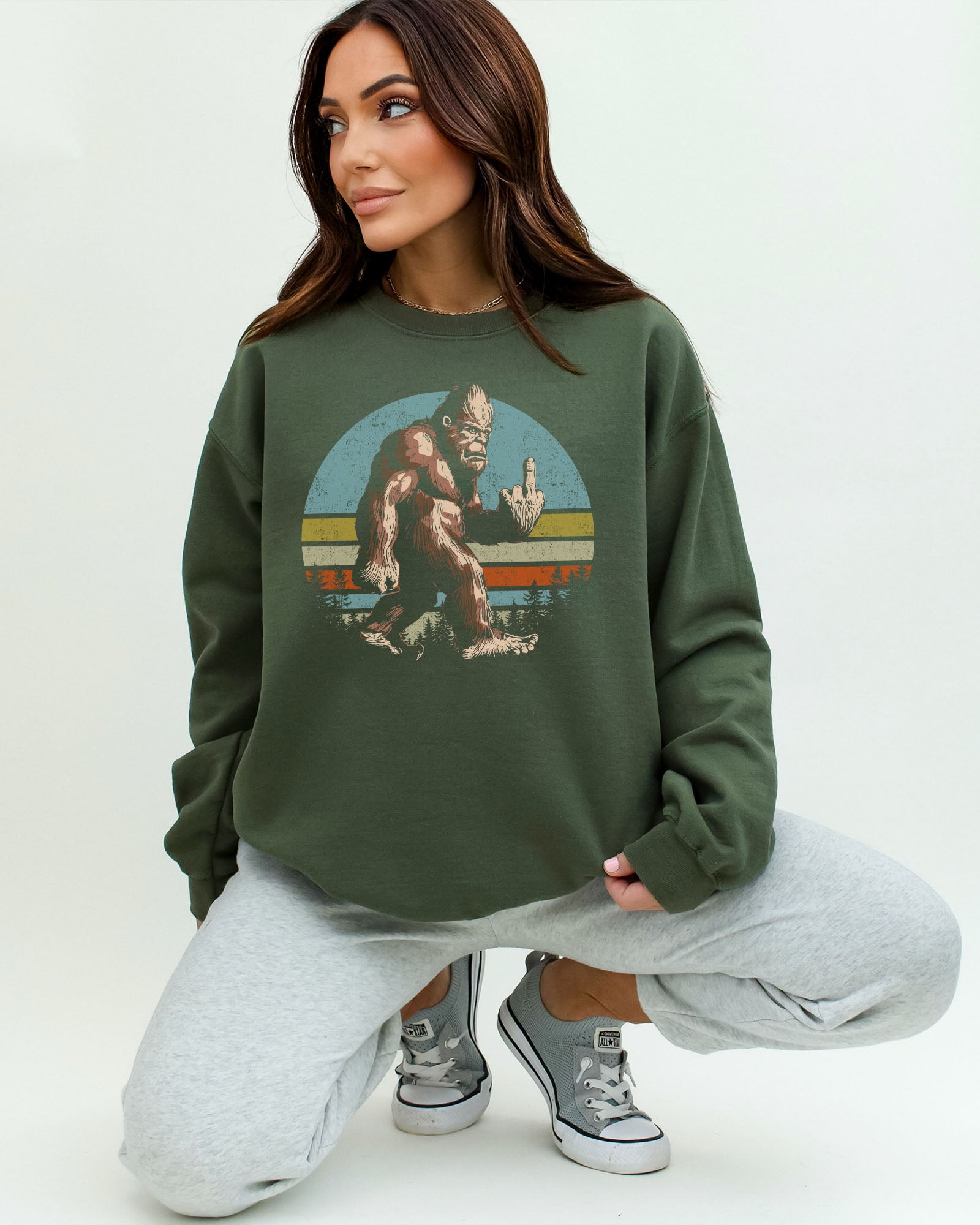 Woman wearing a military green sweatshirt featuring a funny retro-style Bigfoot showing the middle finger graphic.