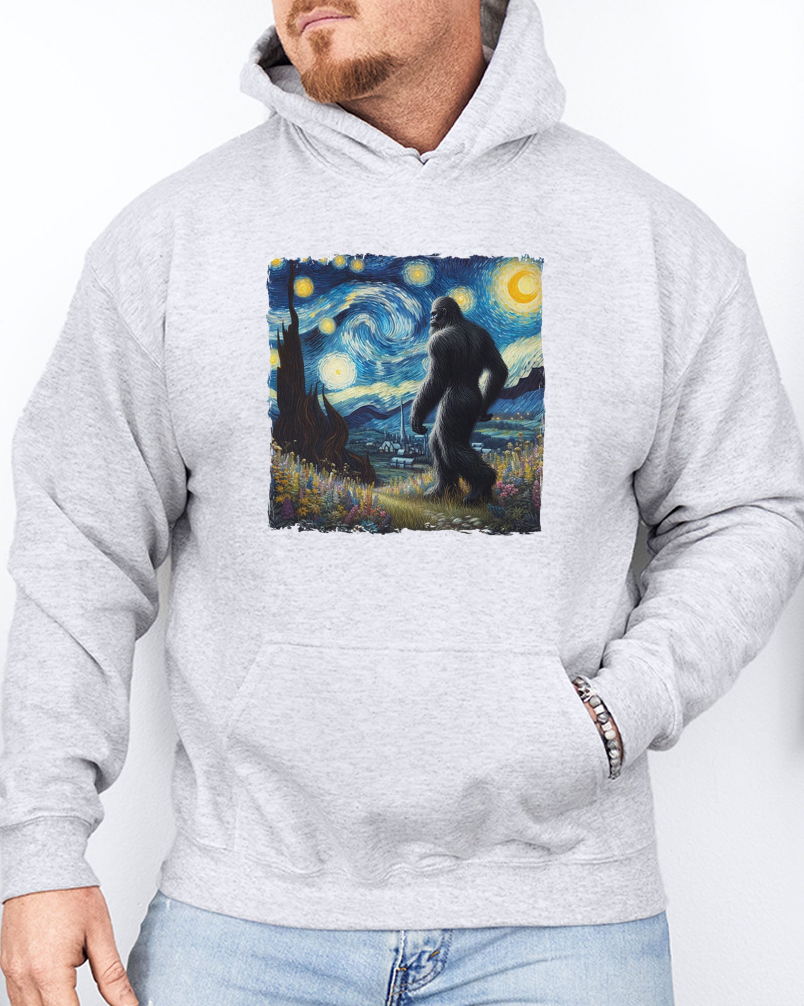 Man wearing an ash gray hoodie featuring a Bigfoot walking through a starry night forest graphic.
