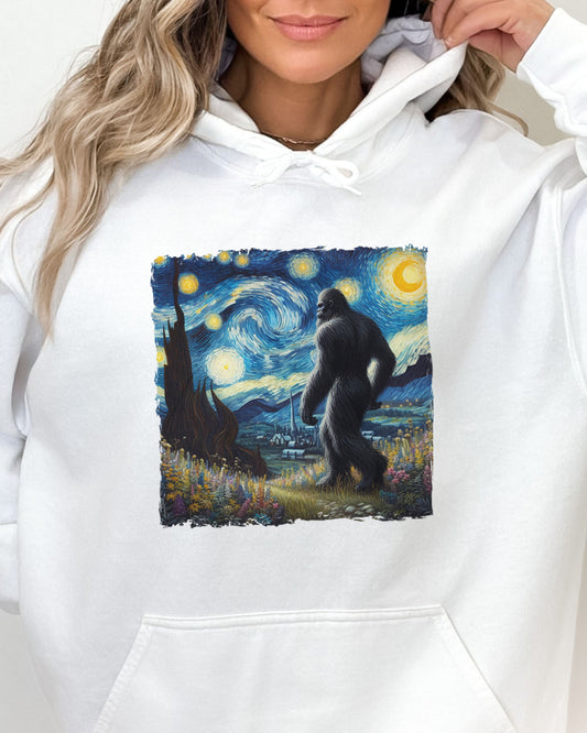 Close-up of a white hoodie featuring a Bigfoot walking through a starry night forest graphic.
