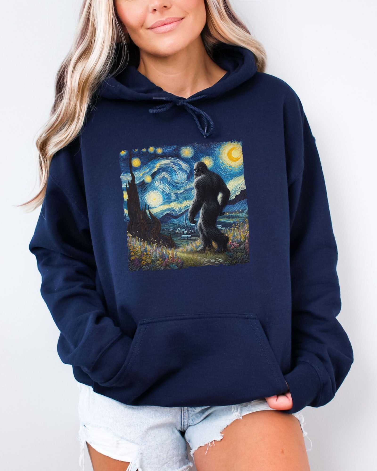 Woman wearing a navy blue hoodie featuring a Bigfoot walking through a starry night forest graphic.