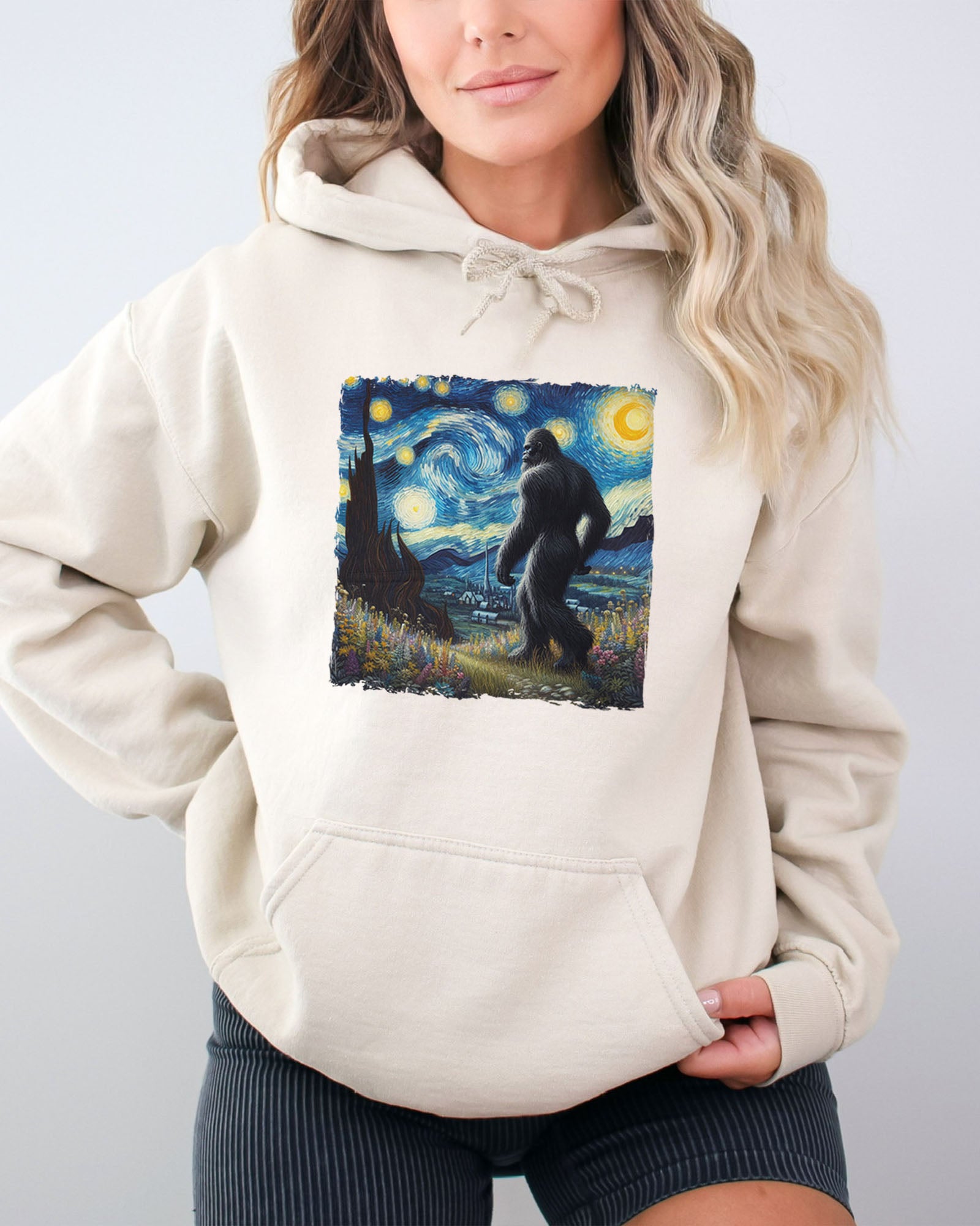 Woman wearing a sand beige hoodie featuring a Bigfoot walking through a starry night forest graphic.