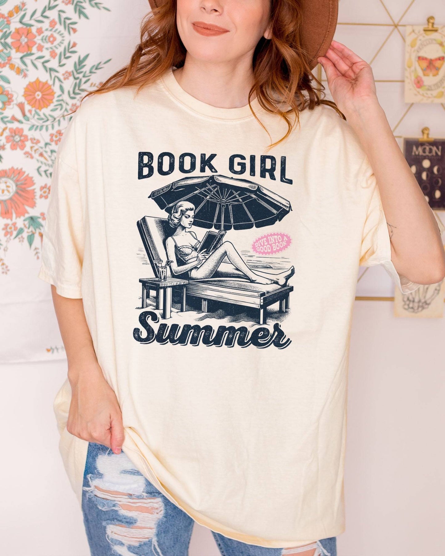 Woman wearing ivory "Book Girl Summer" shirt with retro graphic of a woman on a beach lounger reading a book.