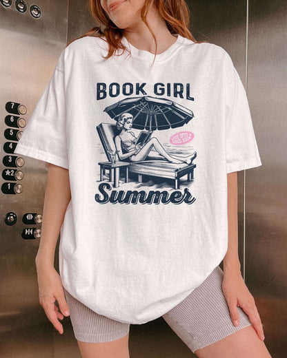 Woman wearing white "Book Girl Summer" shirt with retro graphic of a woman on a beach lounger reading a book.