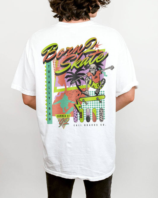 Man wearing a white “Born 2 Skate” shirt with a retro 90s-style graphic of a skeleton skateboarding and bold skateboard designs.