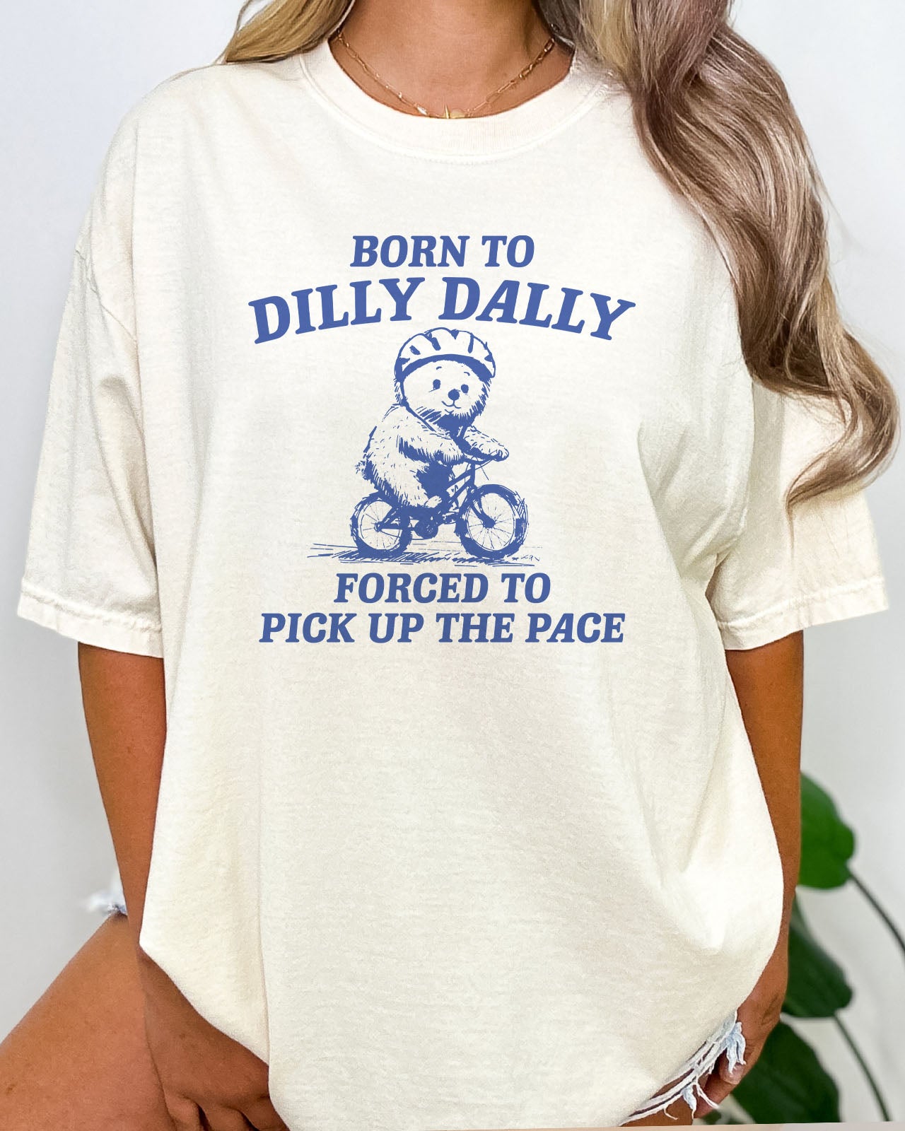 Woman wearing Born To Dilly Dally Shirt in natural beige color showing a retro bear graphic and the quote "Born To Dilly Dally, Forced To Pick Up The Pace"